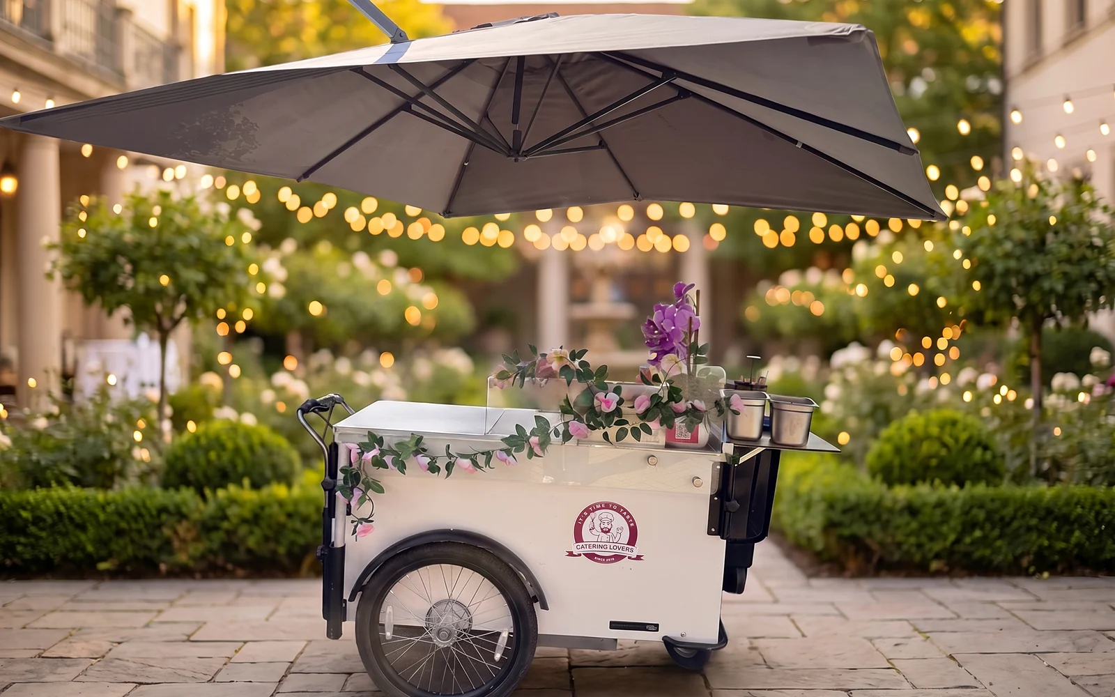 HappyBike Small: weißer Pushcart mit Rosen-Deko unter dem Schirm, abends im Innenhof mit Lichterketten