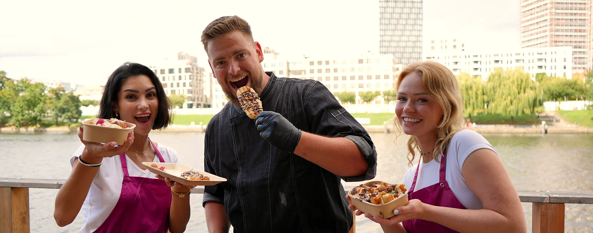 Live-Cooking in Berlin: Koch beißt in einen MilkyStick, zwei Crew-Kolleginnen lachen mit Waffelschalen — dahinter Spree und Berliner Skyline.