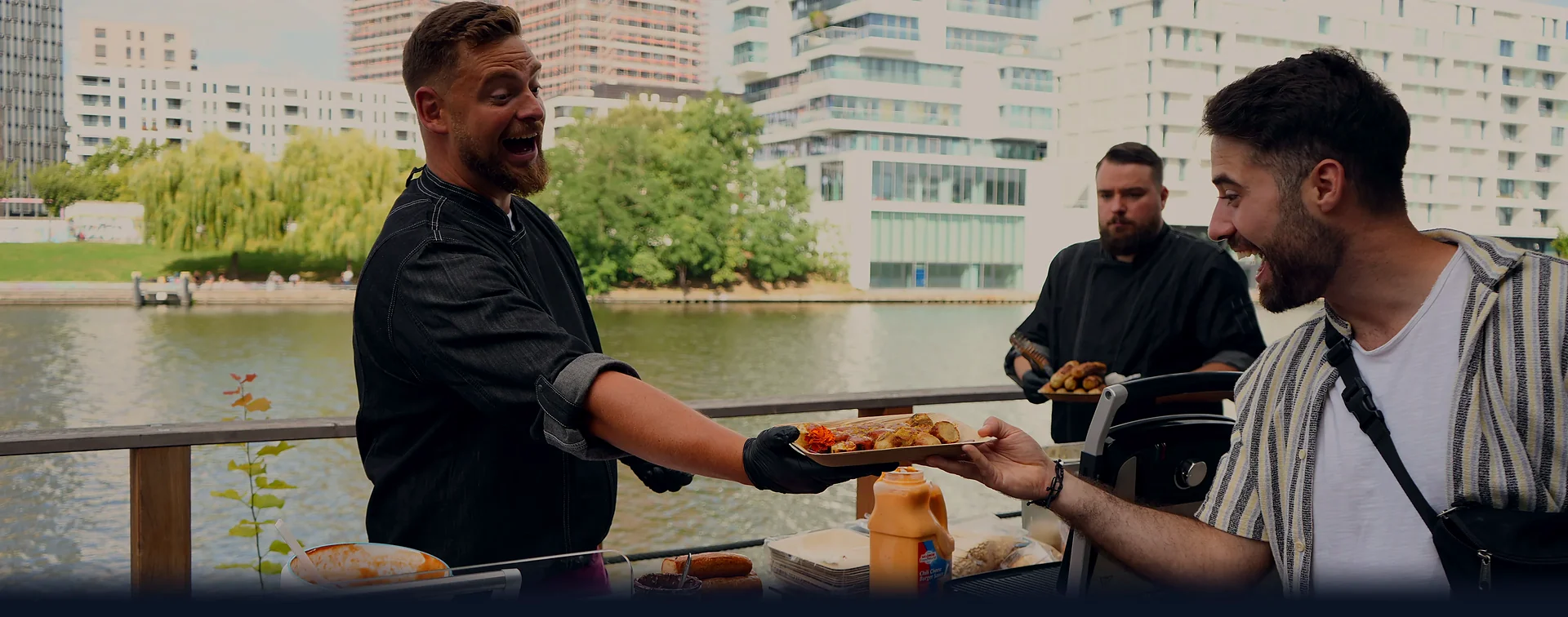 Firmenfeier am Wasser: CateringLovers-Koch übergibt frisch zubereitete Leckereien lachend an einen Gast.