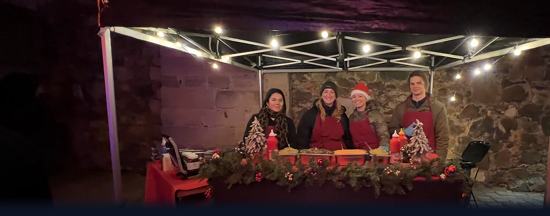 CateringLovers-Crew in roten Schürzen, eine mit Weihnachtsmütze, an einem beleuchteten Weihnachtsstand mit Tannengirlande unter dem Pavillon am Abend