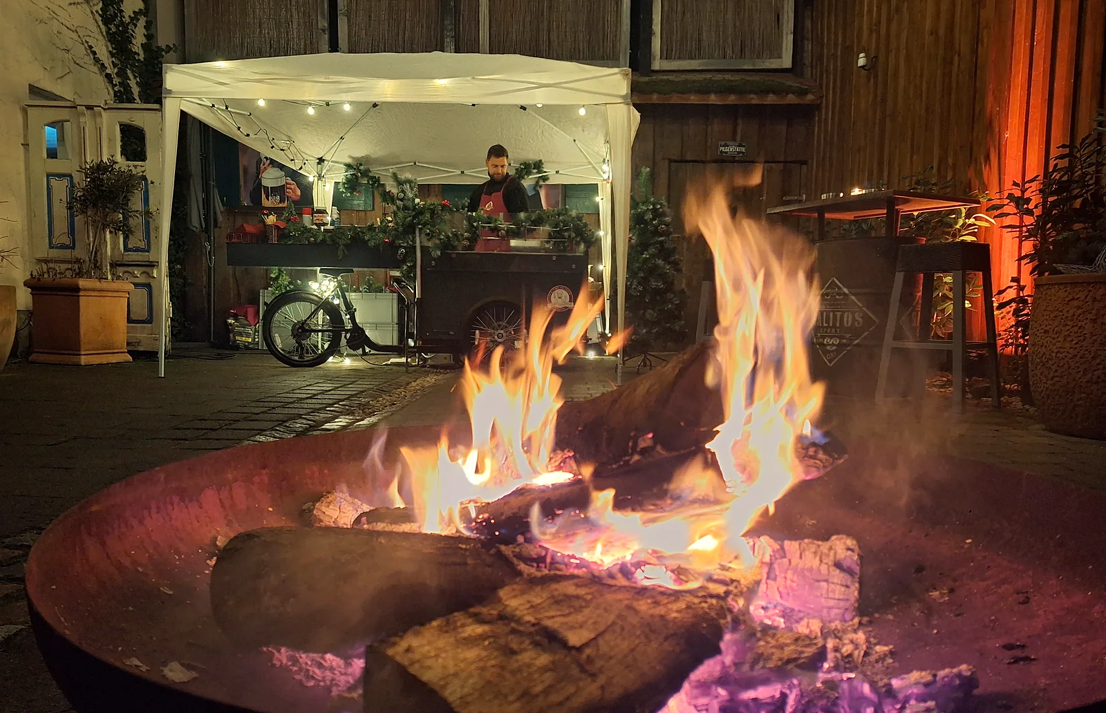 Weihnachtsmarkt im Innenhof am Abend: vorn eine lodernde Feuerschale, dahinter das geschmückte HappyBike unter dem beleuchteten Pavillon
