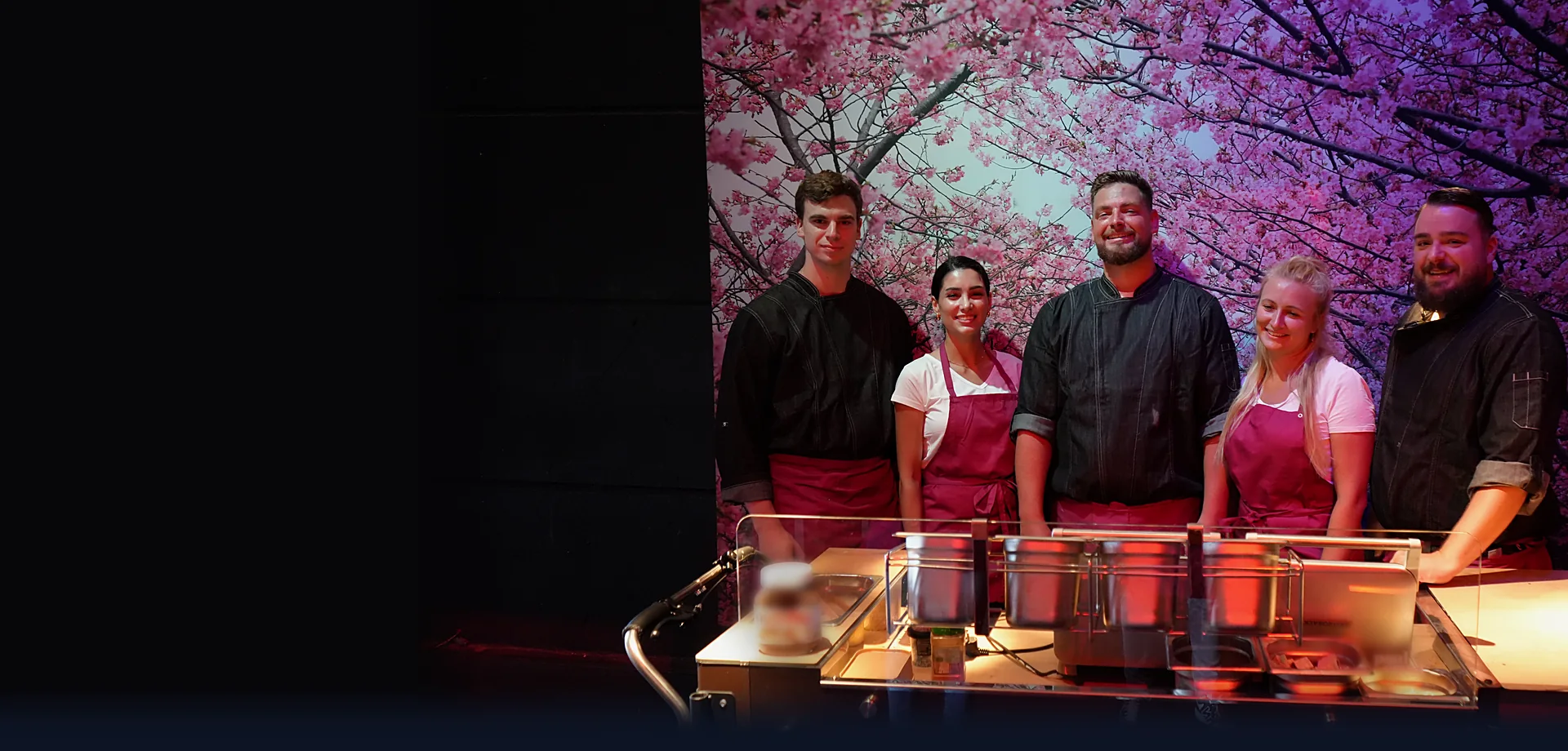Fünf Crew-Mitglieder von CateringLovers an ihrer Live-Cooking-Station vor einer Wand aus Kirschblüten