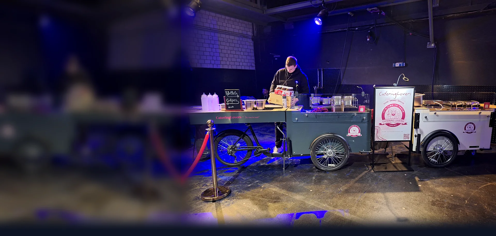Zwei HappyBikes von CateringLovers mit Waffel- und Crêpes-Station in einer dunklen Eventhalle, ein Crew-Mitglied bereitet die Ausgabe vor