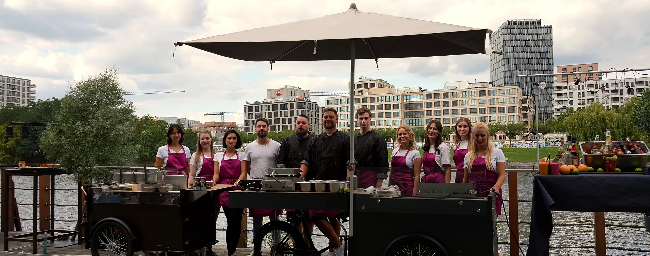 CateringLovers-Crew am HappyBike-Verbund unter dem Schirm, dahinter Fluss und Skyline