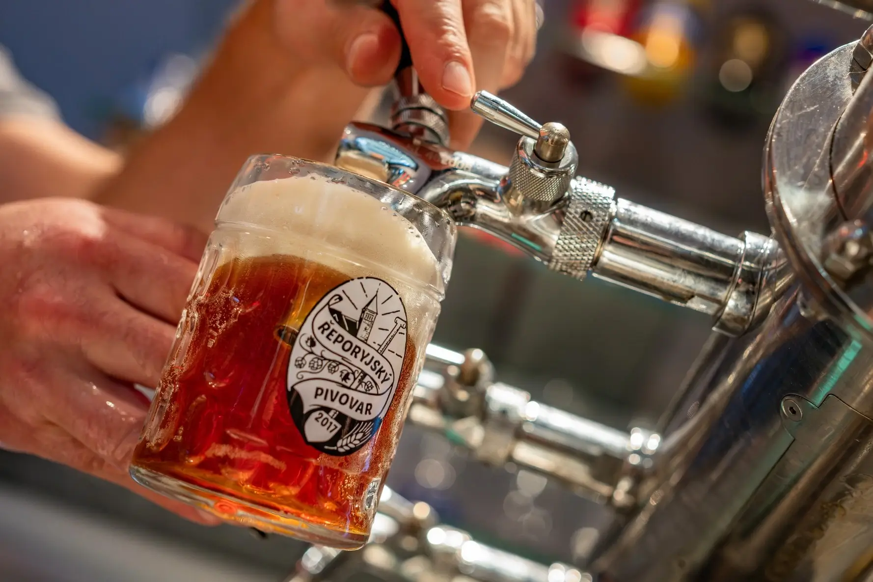 Hand holding a glass mug of amber beer being poured from a shiny metal tap with a 'RĚPORYJSKÝ PIVOVAR' logo.