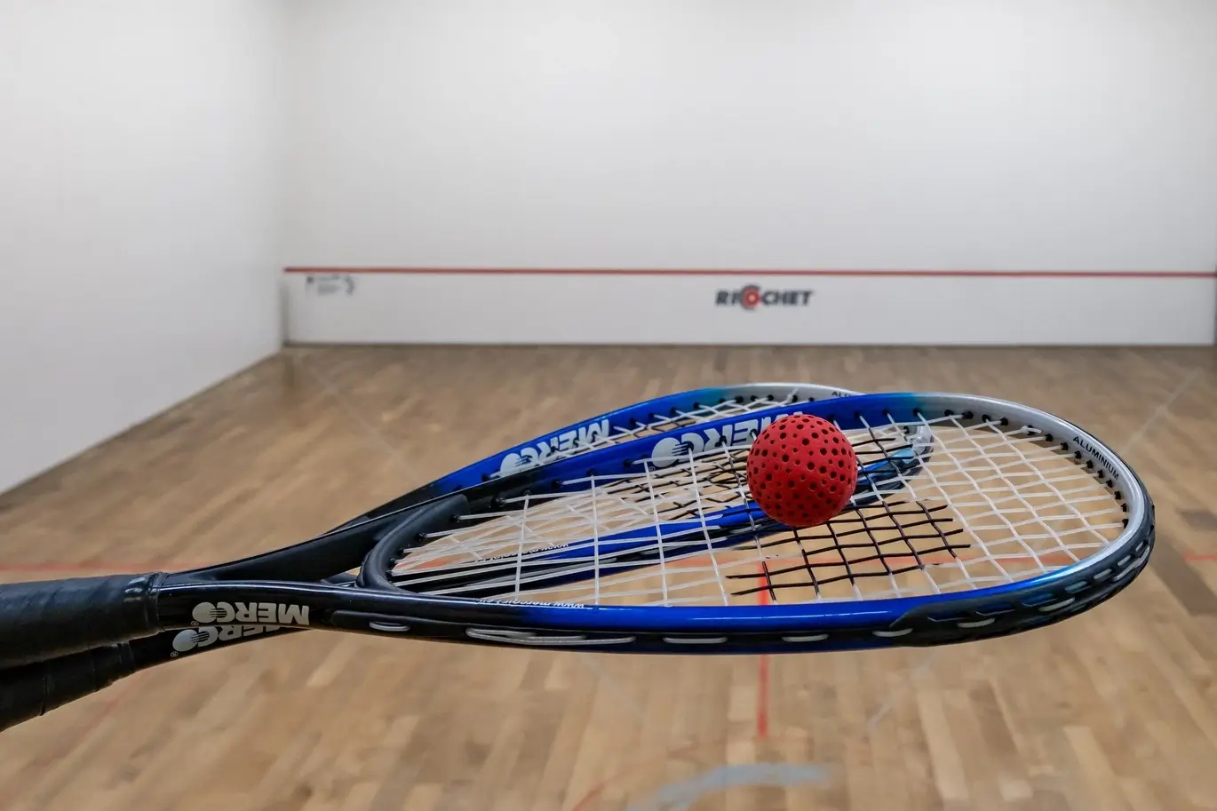Two blue racquets with a red perforated ball resting on top inside a squash court.