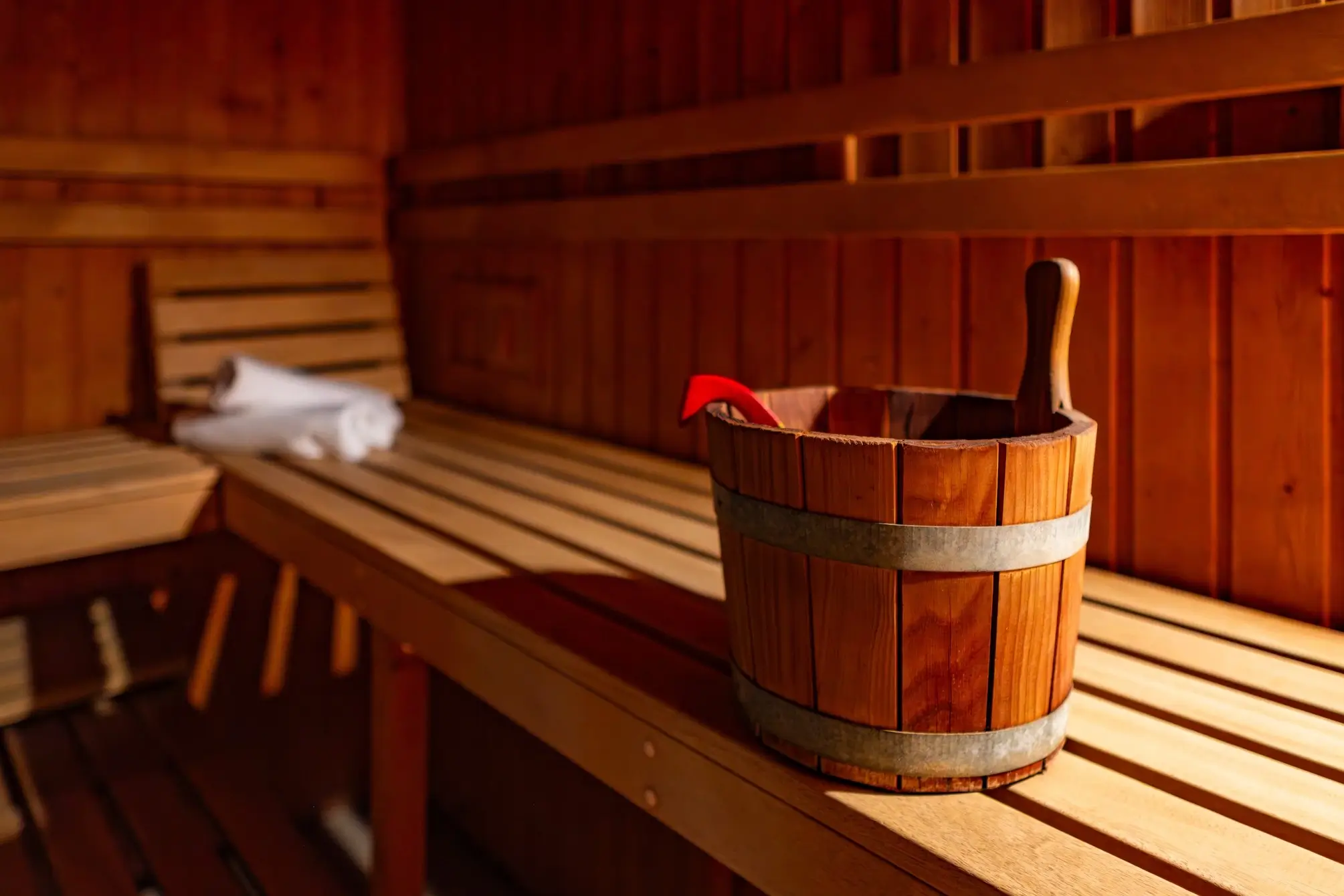 Wooden bucket with a ladle inside placed on a wooden bench in a sauna room with rolled white towels in the background.
