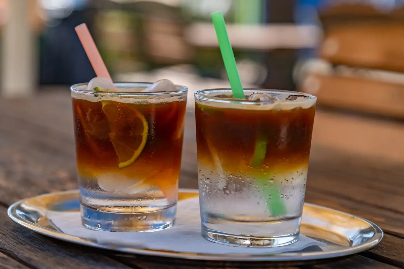 Two glasses of iced layered coffee with lemon slices and colorful straws on a metal tray.