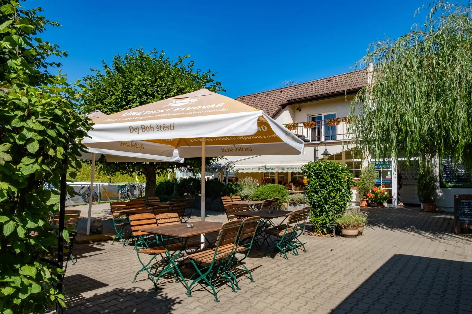 Outdoor patio seating area with wooden tables and chairs under beige umbrellas at a restaurant with greenery and a building in the background.