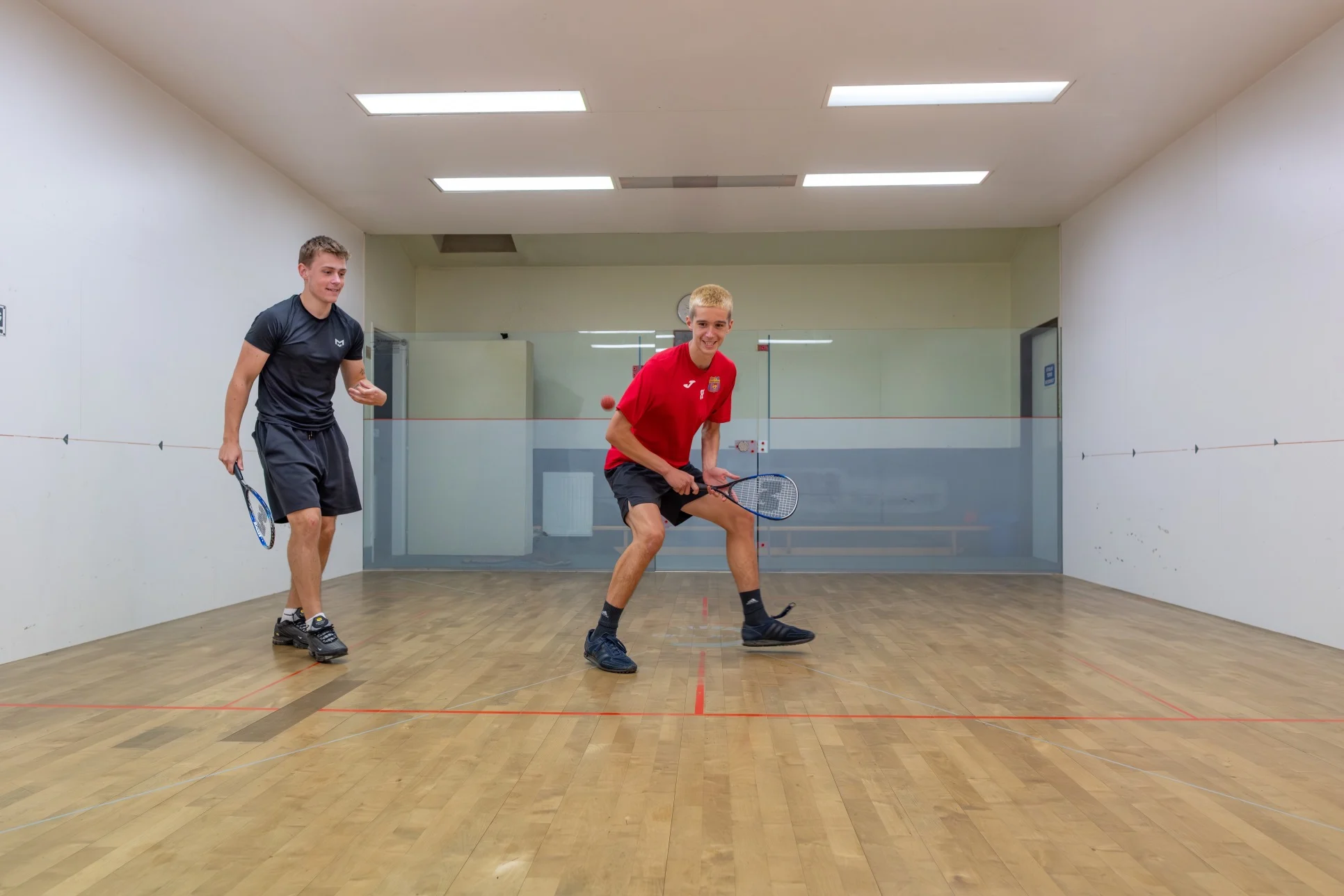 Two young men playing squash in an indoor court, one in a red shirt preparing to hit the ball and the other in a black shirt ready with a racket.