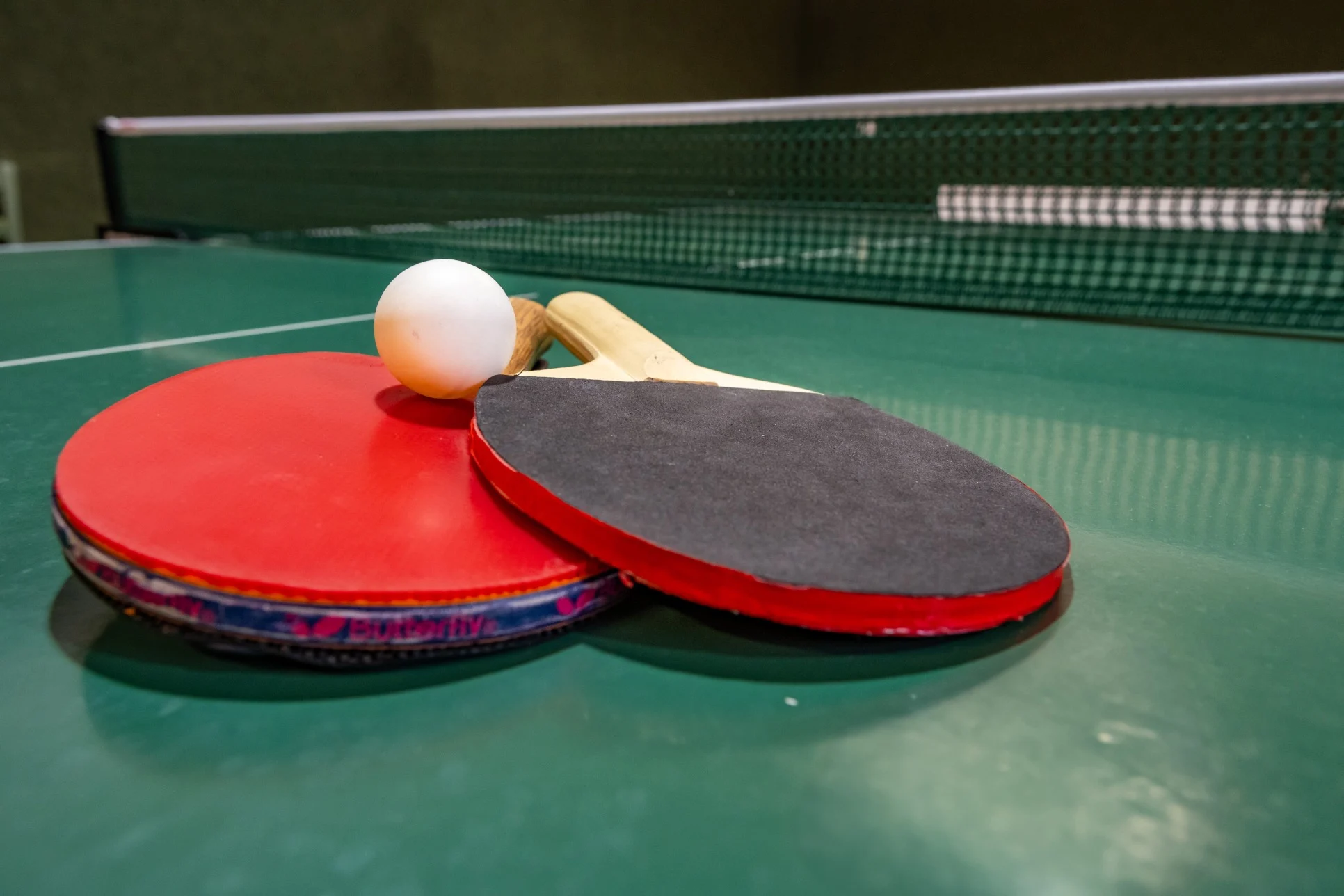 Two table tennis paddles, one red and one black, with a white ping pong ball resting on a green table tennis table.