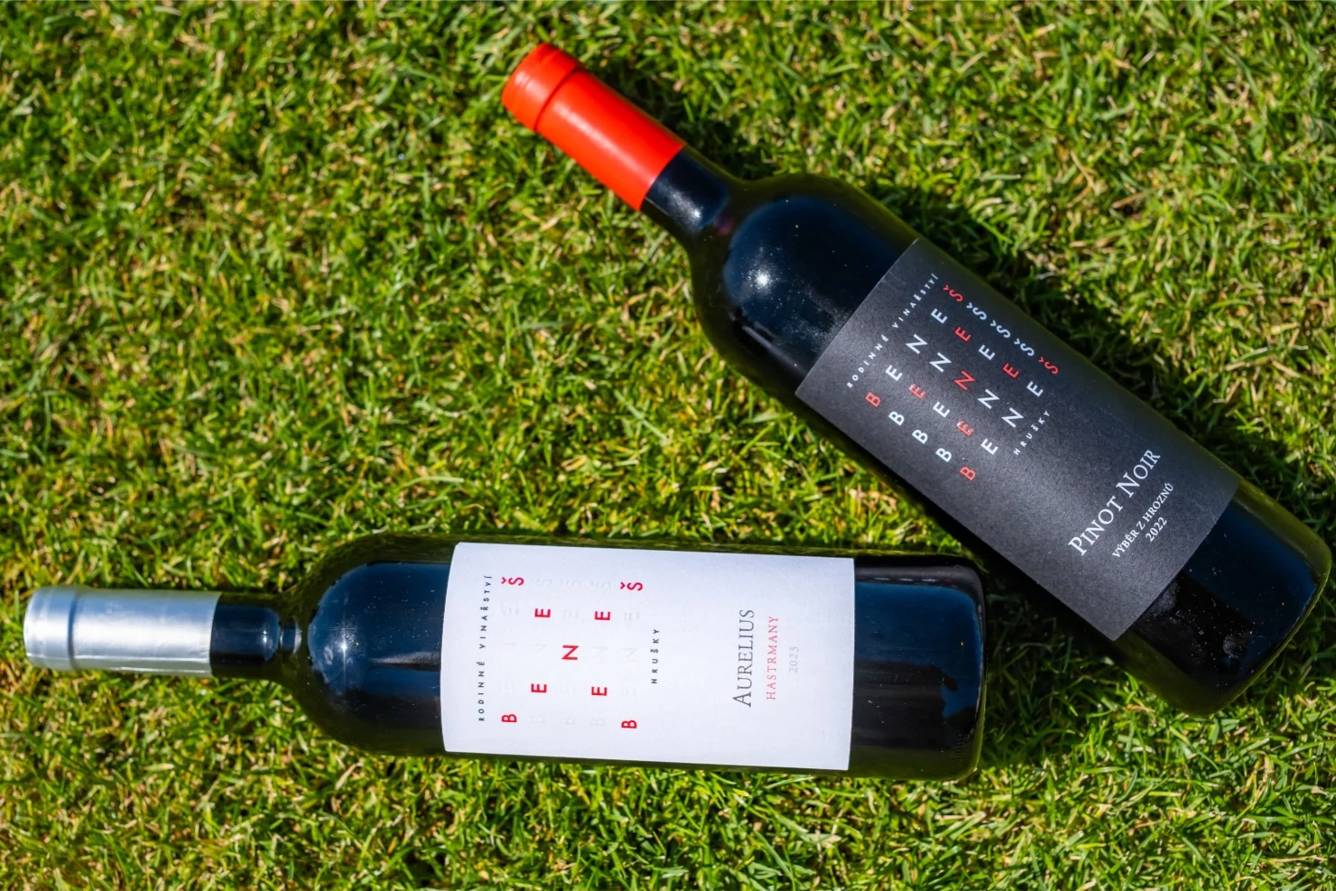 Two bottles of red wine lying on grass, one with a red cap labeled Pinot Noir and the other with a silver cap labeled Aurelius.