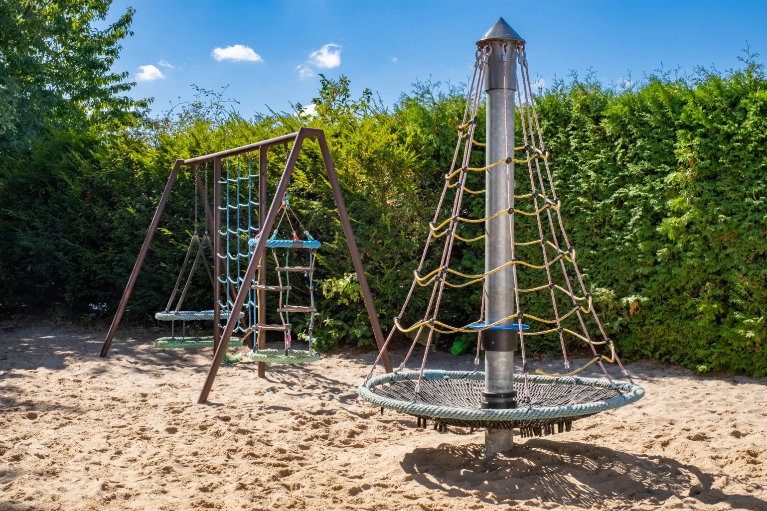 Playground with a round rope climbing structure and a swing set on sandy ground, surrounded by green bushes under a blue sky.