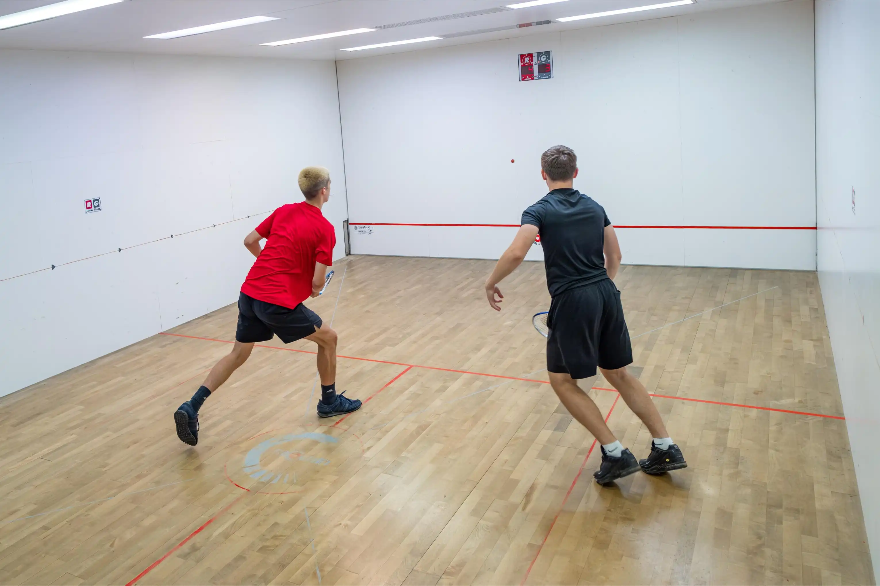 Two men playing squash in an indoor court, one wearing a red shirt and the other in black.