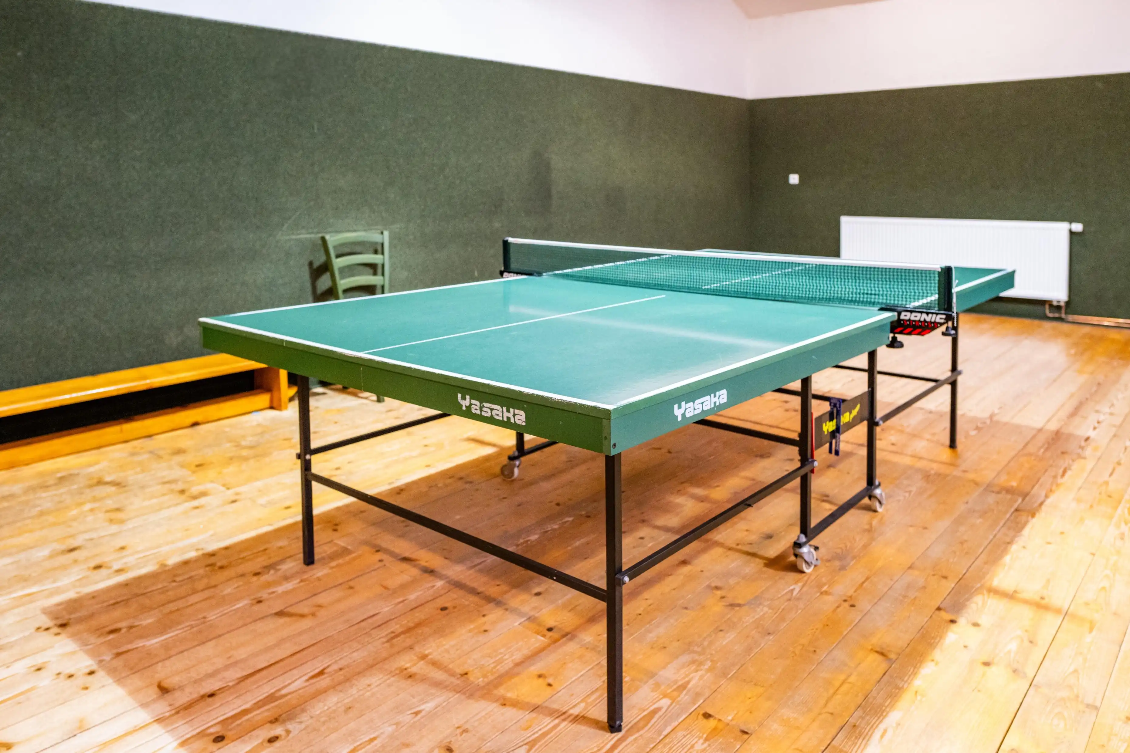 Green Yasaka table tennis table with a Donic net in a room with wooden floors and green walls.