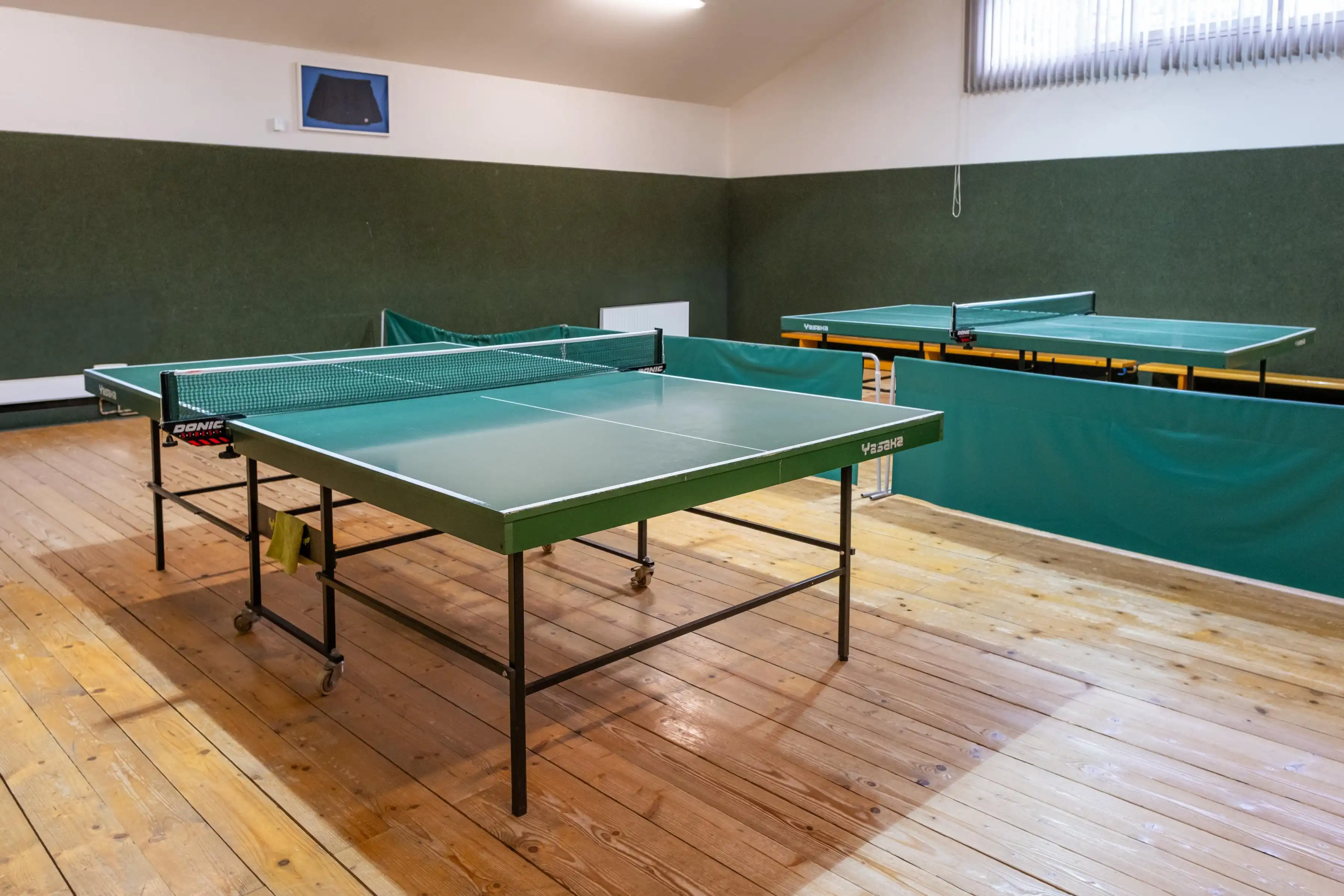 Indoor room with two green table tennis tables on wooden floor and green padded walls.