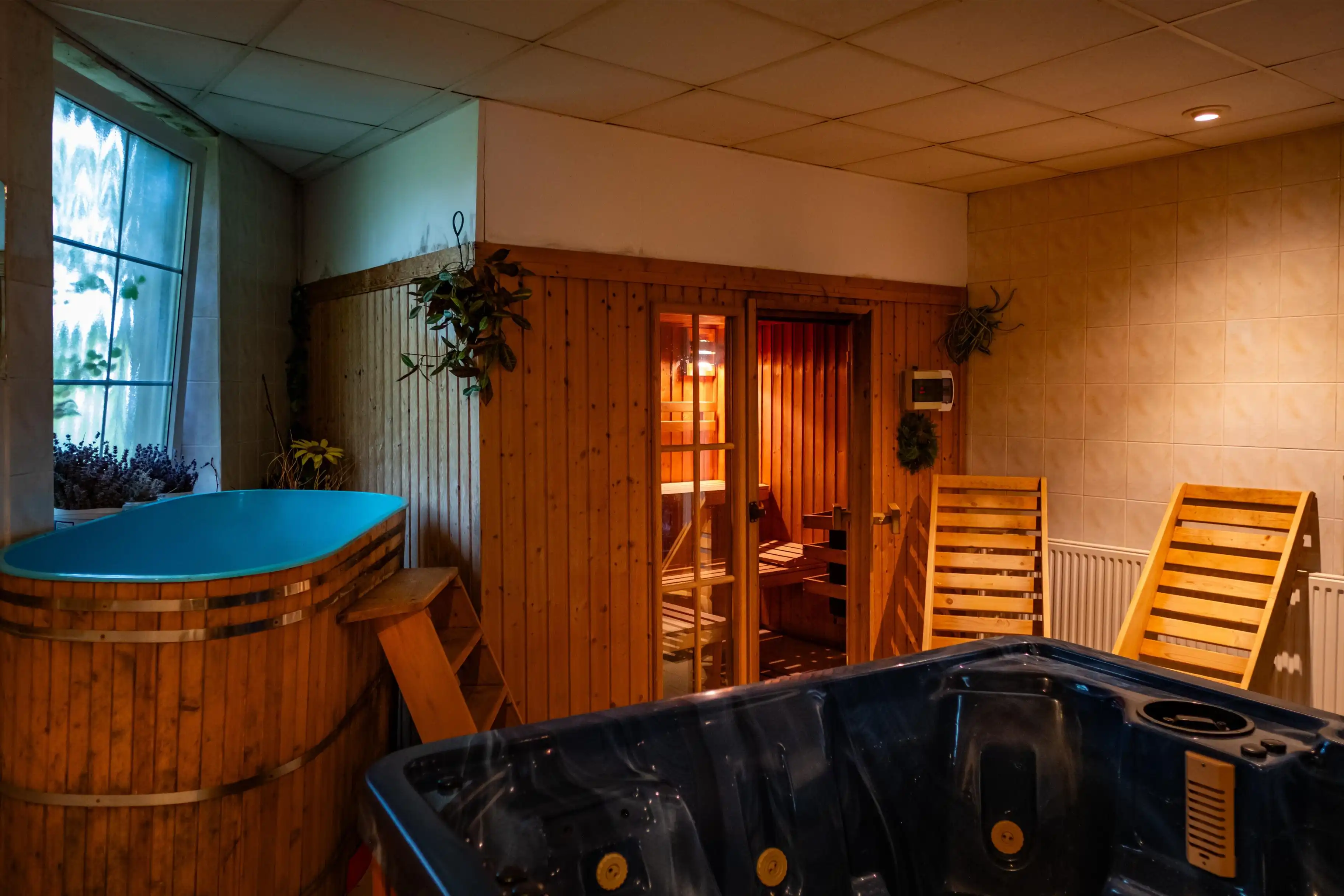 Indoor spa room with a wooden hot tub, a blue jacuzzi, two wooden lounge chairs, and a lit sauna with wooden walls and a glass door.