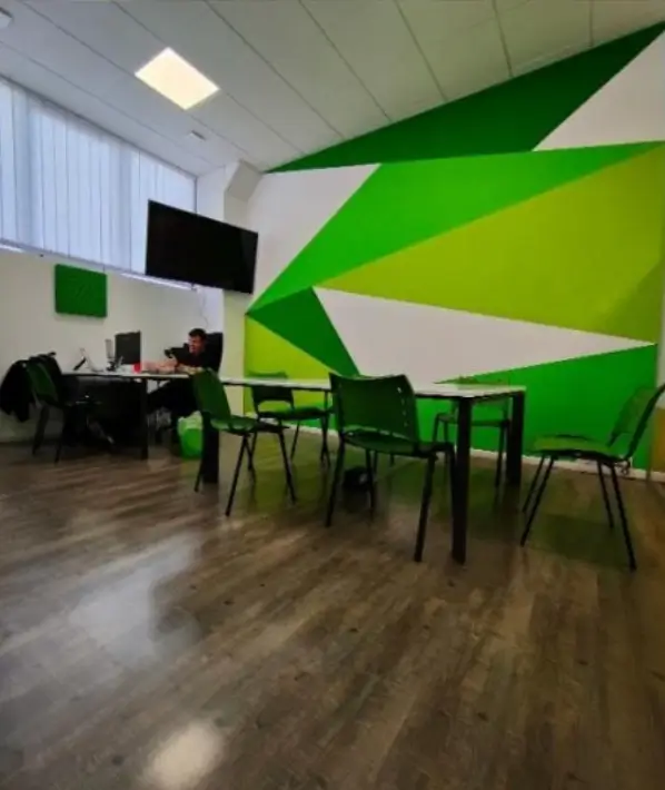 Sala riunioni con tavoli e sedie verdi, una parete decorata con forme geometriche verdi e bianche, e una persona seduta al computer.