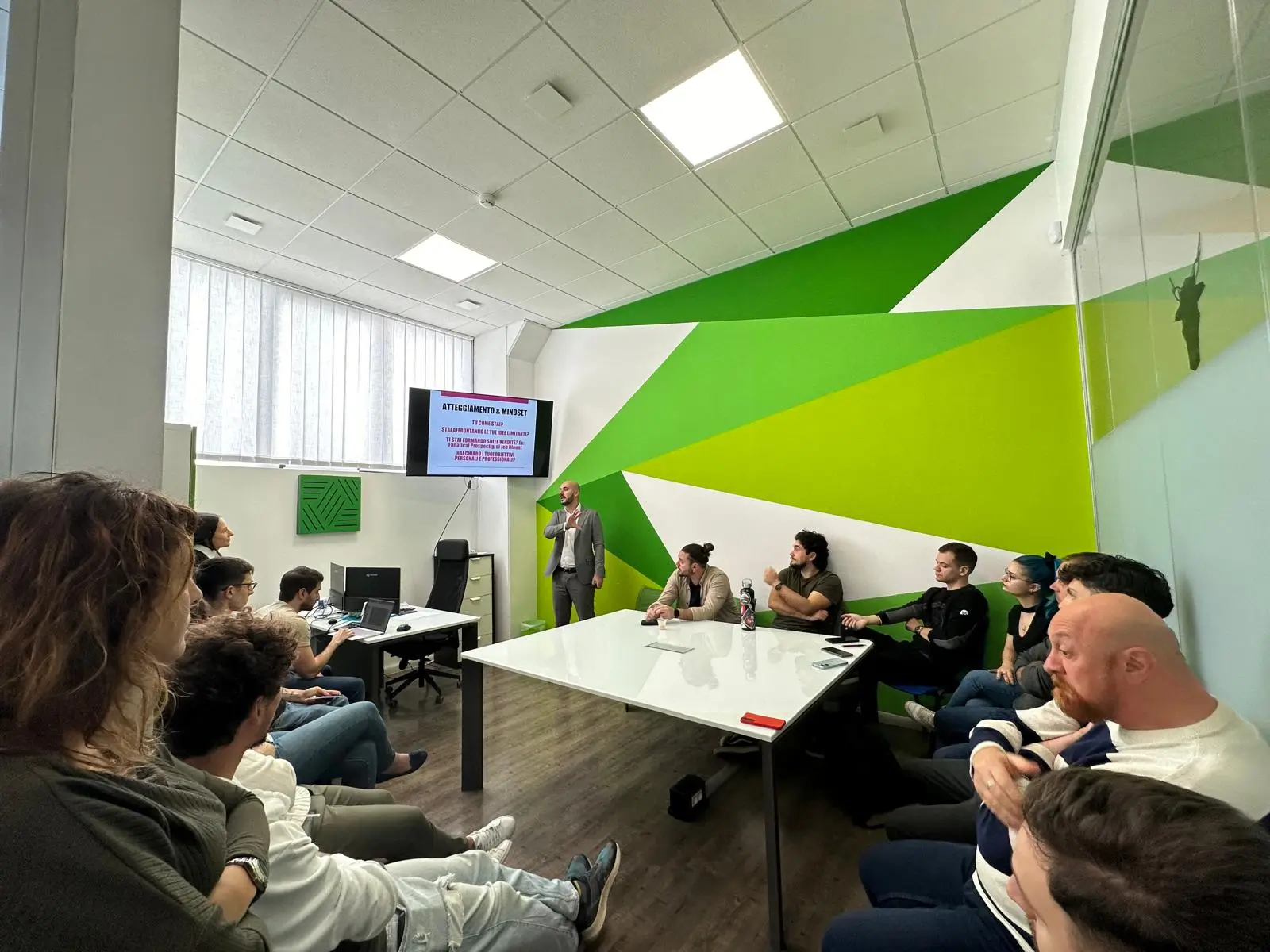 Una persona in piedi che tiene una presentazione davanti a un gruppo di persone sedute in una sala riunioni con pareti dipinte di verde e bianco.