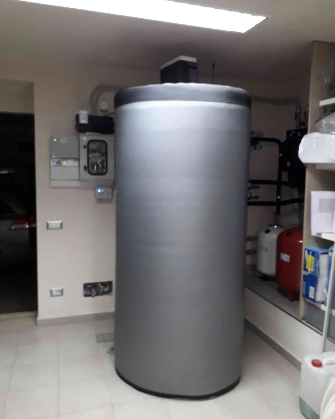 Serbatoio cilindrico grigio all'interno di una stanza tecnica con tubazioni e scatole elettriche.