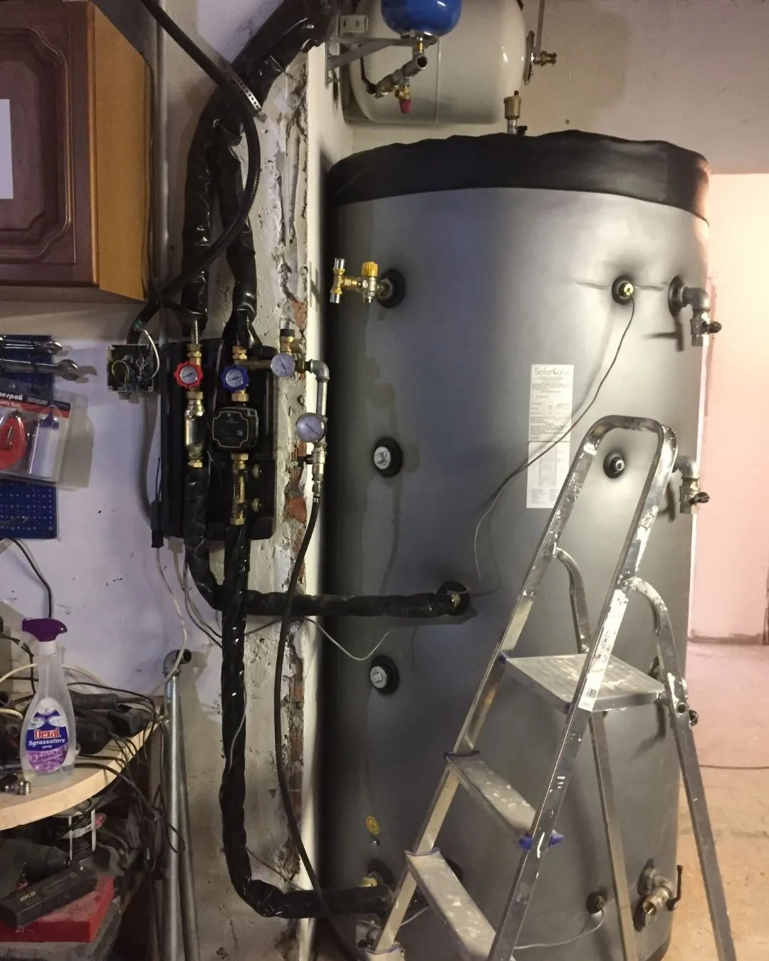 Serbatoio d'acqua con tubazioni isolate e manometri in locale tecnico con scala in metallo davanti.