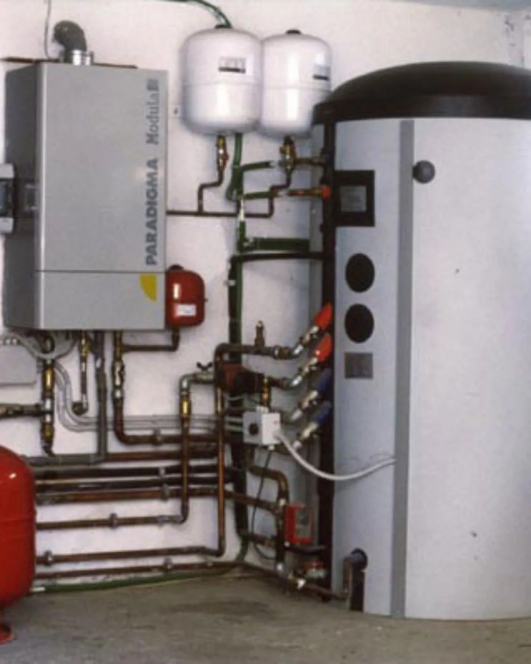 Sistema di riscaldamento con caldaia Paradigma Modella, serbatoi bianchi e tanica di accumulo grigia con tubazioni collegate.