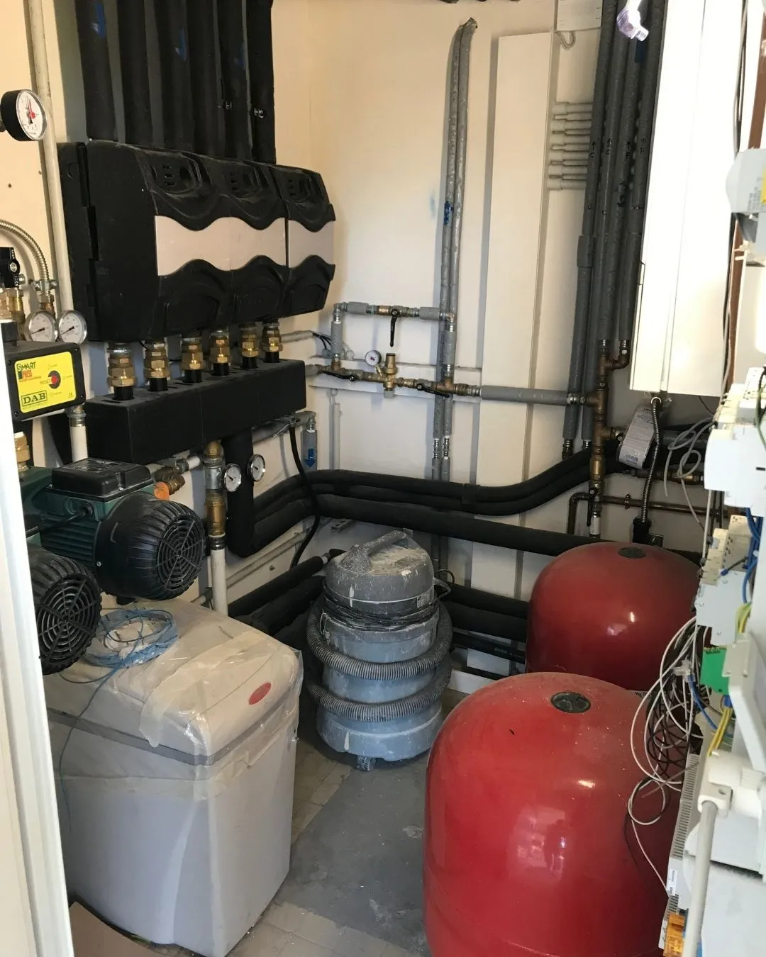 Locale tecnico con tubi isolati, caldaia, due serbatoi rossi, pompa e aspirapolvere industriale.