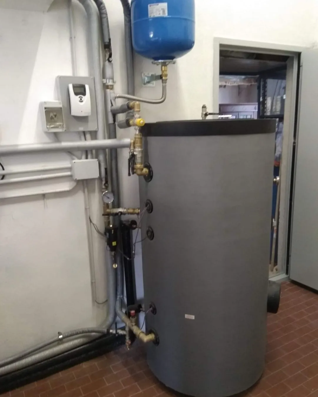 Serbatoio cilindrico grigio con tubazioni e valvole in un locale tecnico con pavimento in mattoni.