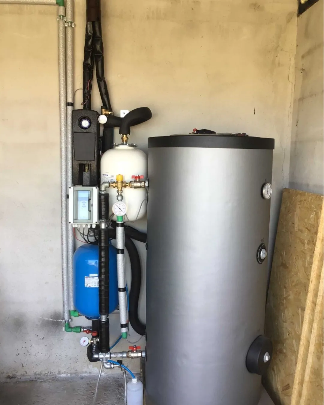 Impianto di riscaldamento domestico con serbatoio cilindrico grigio, tubazioni isolate e serbatoio di espansione blu e bianco montati su una parete di cemento.