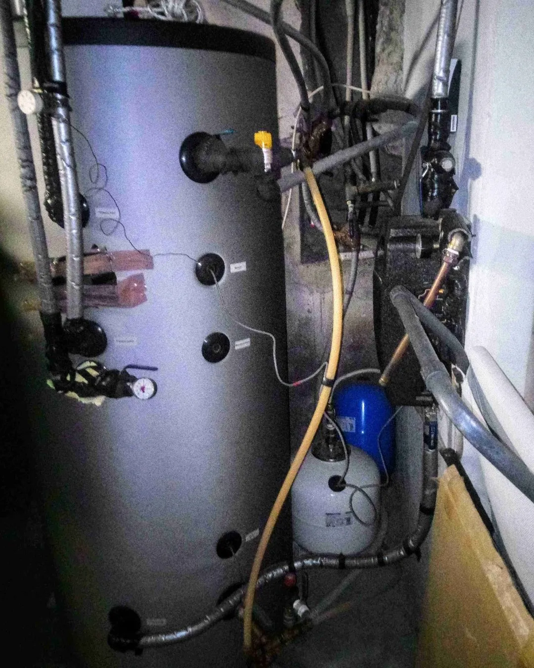 Impianto di riscaldamento con serbatoio cilindrico grigio, tubi isolati e valvole in una stanza tecnica.
