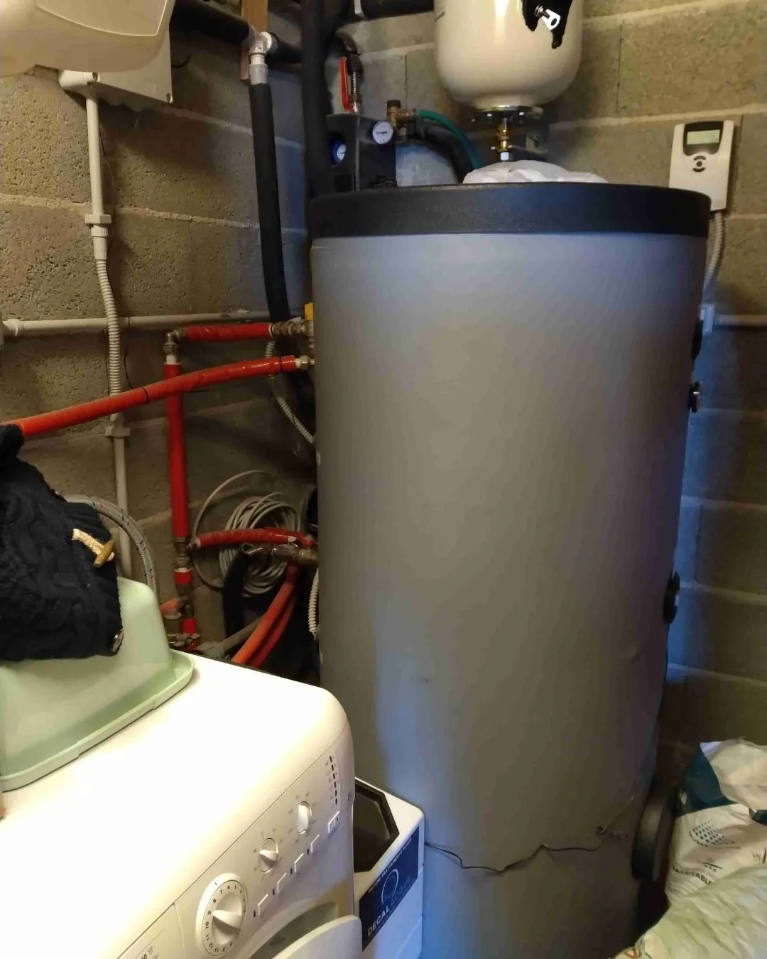 Caldaia grigia con tubi rossi e neri in un locale tecnico con pareti in cemento e una lavatrice bianca.