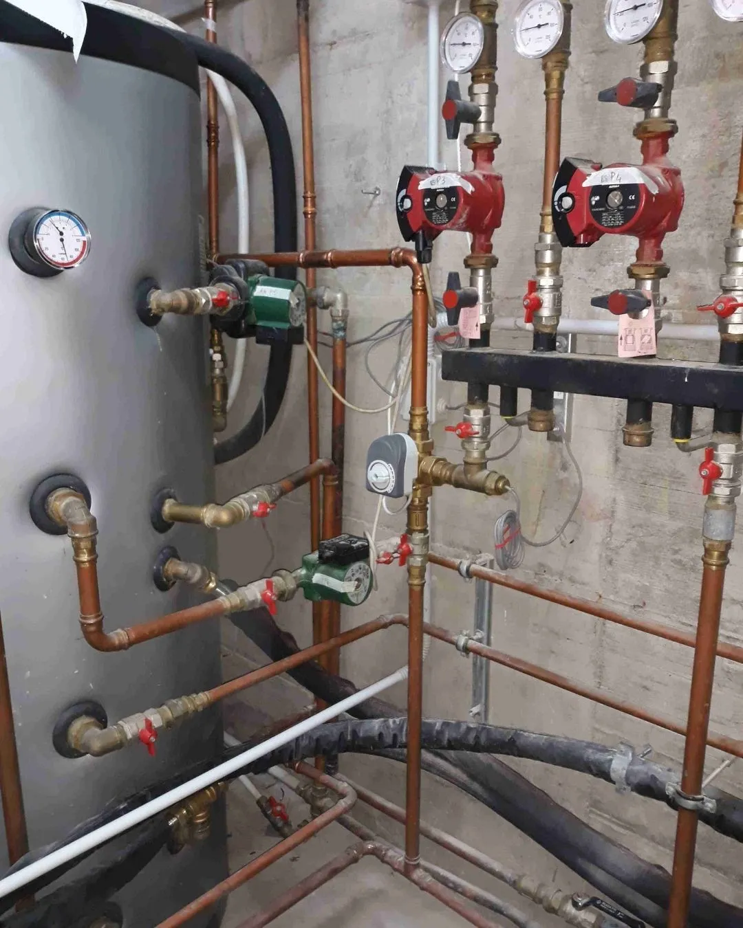 Impianto di tubature e valvole in un locale tecnico con manometri e pompe di circolazione rosse e verdi.