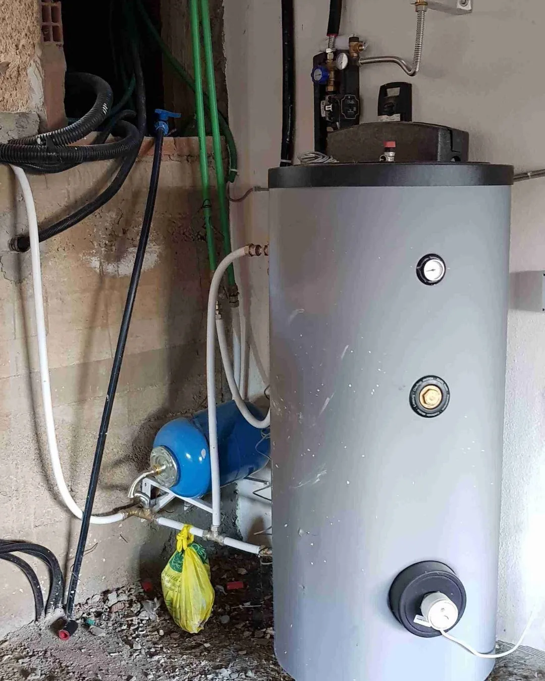 Caldaia grigia cilindrica con tubi e componenti collegati in una stanza con pareti di cemento grezzo.