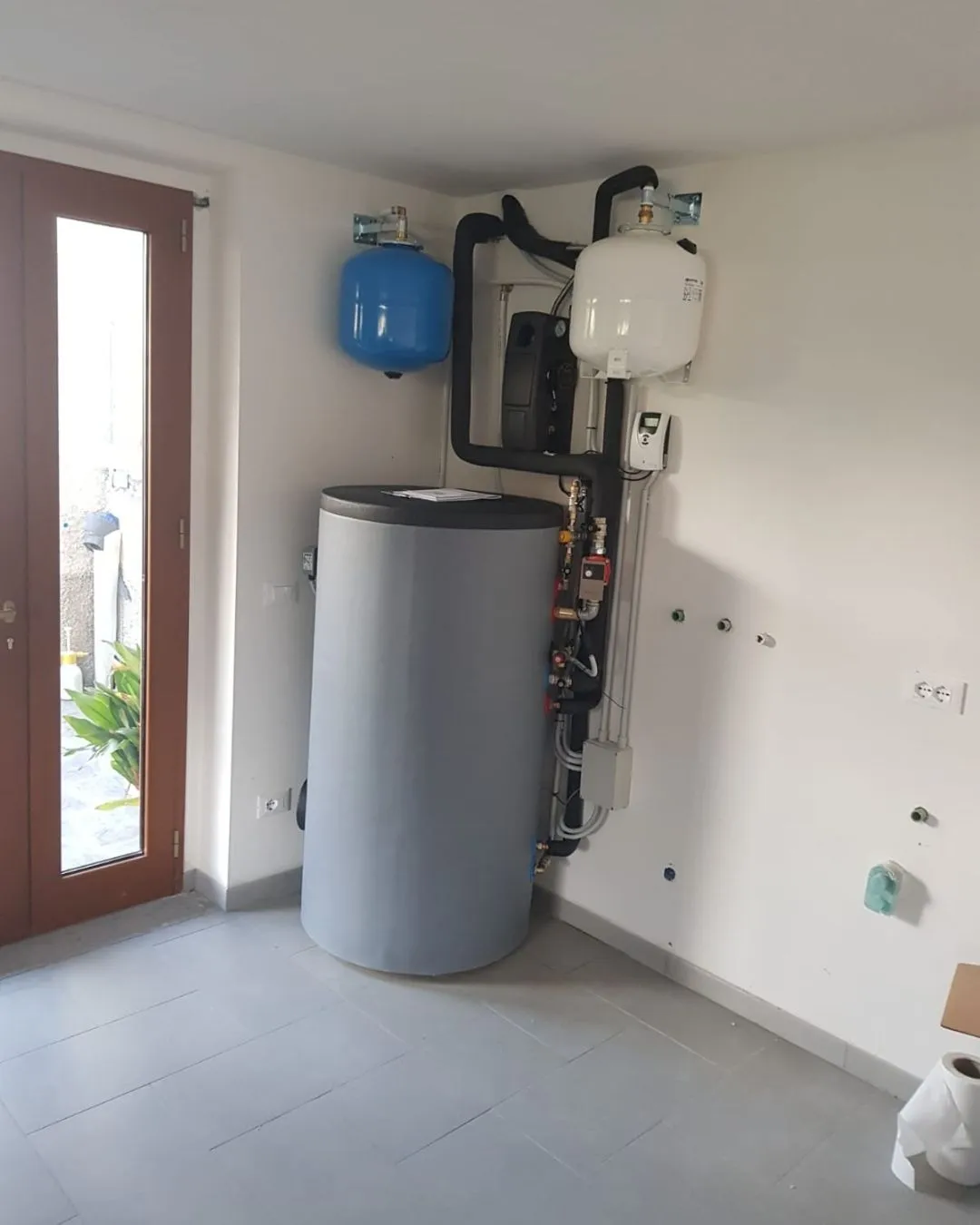 Locale tecnico con caldaia cilindrica grigia, due serbatoi di espansione uno blu e uno bianco montati su parete, e tubature nere.