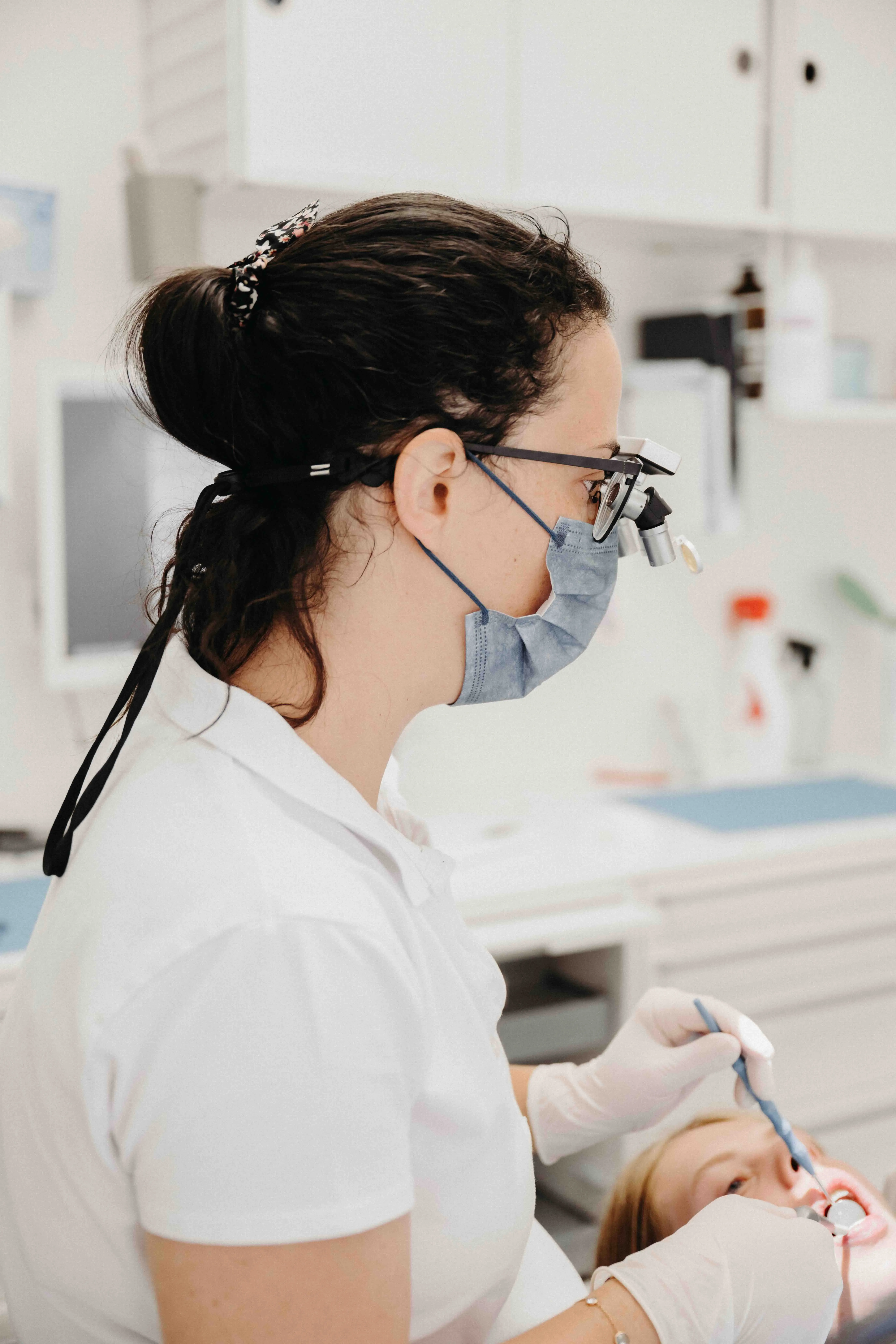 Dentiste en blouse blanche, portant un masque et des lunettes avec loupe, traitant la bouche d'un patient allongé dans un fauteuil dentaire dans un cabinet moderne.