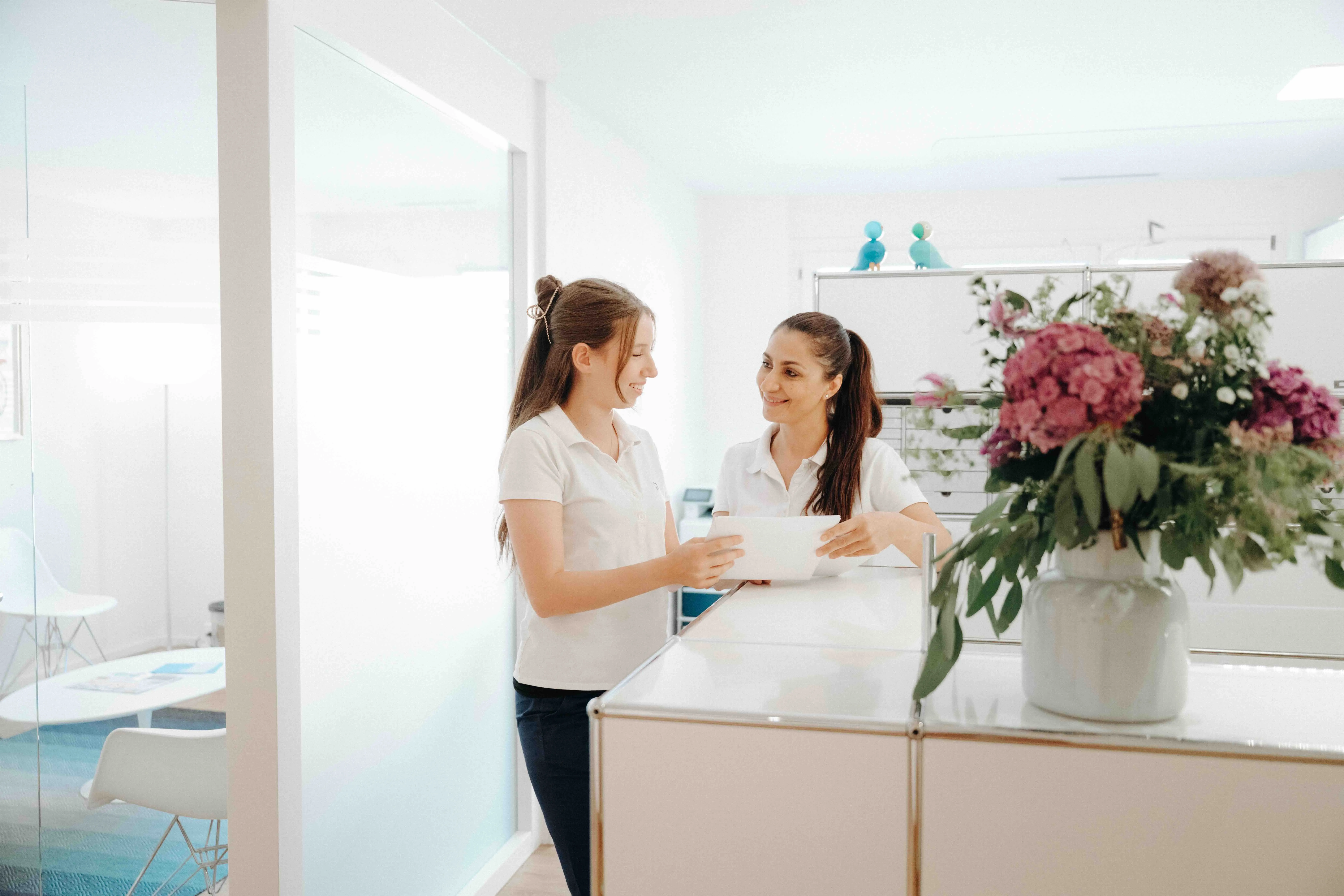 Deux femmes souriantes en chemises blanches discutant à un bureau moderne dans un espace lumineux avec un vase de fleurs violettes sur le comptoir.