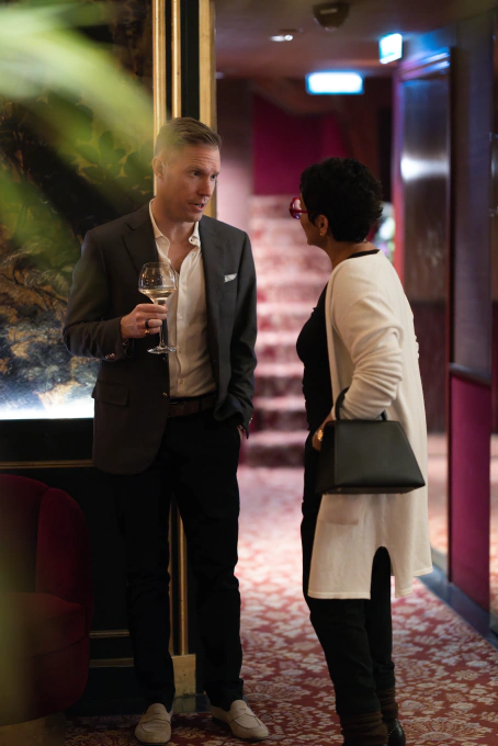 Man in a dark suit holding a wine glass talking to a woman wearing a white cardigan and carrying a black handbag in a warmly lit interior.