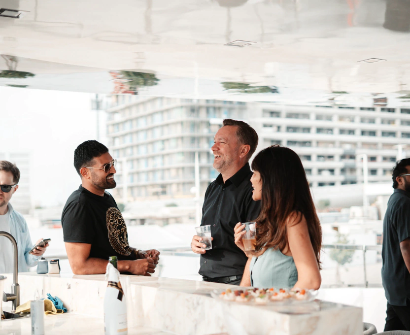 Three people casually chatting and holding drinks at a modern outdoor bar with a cityscape background.