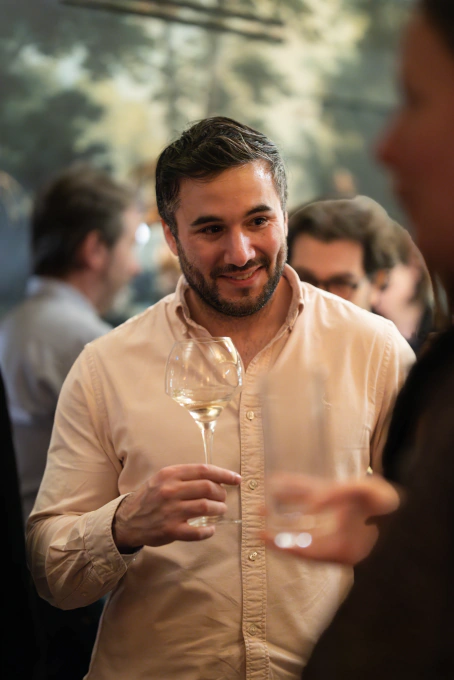 Man with short dark hair and beard wearing a beige shirt, holding a wine glass and smiling during a social gathering.