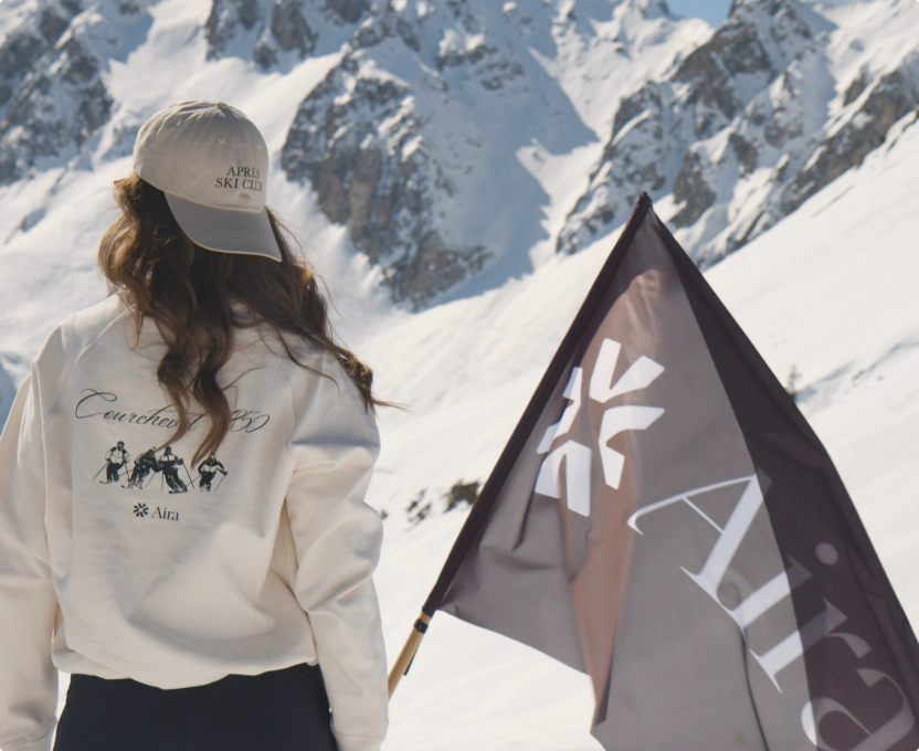 Person in white sweatshirt and beige cap holding a black flag with mountain snow background.