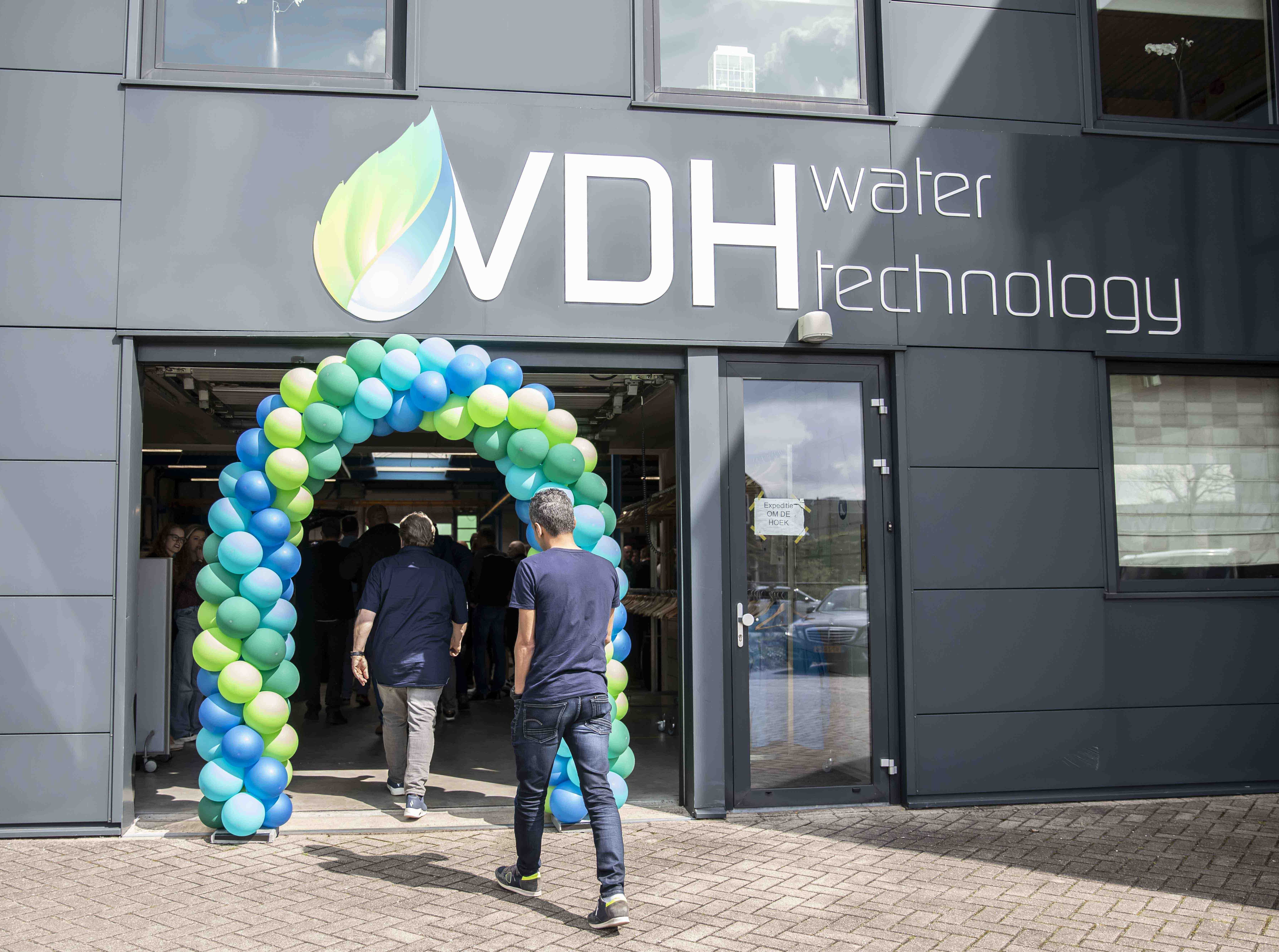 People entering a building under a blue and green balloon arch with a sign reading VDH water technology on the wall above.