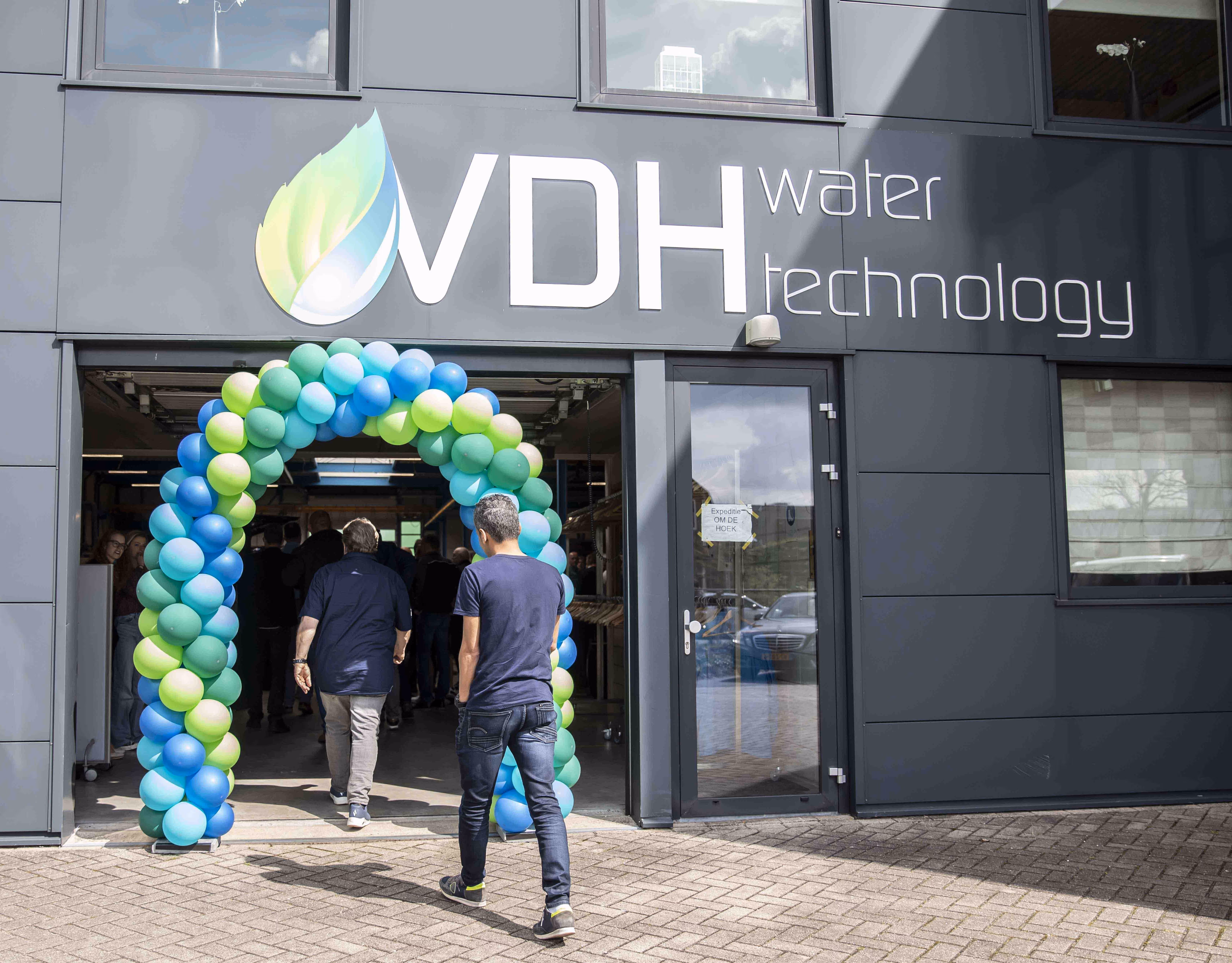 People entering a building under a blue and green balloon arch with a sign reading VDH water technology on the wall above.