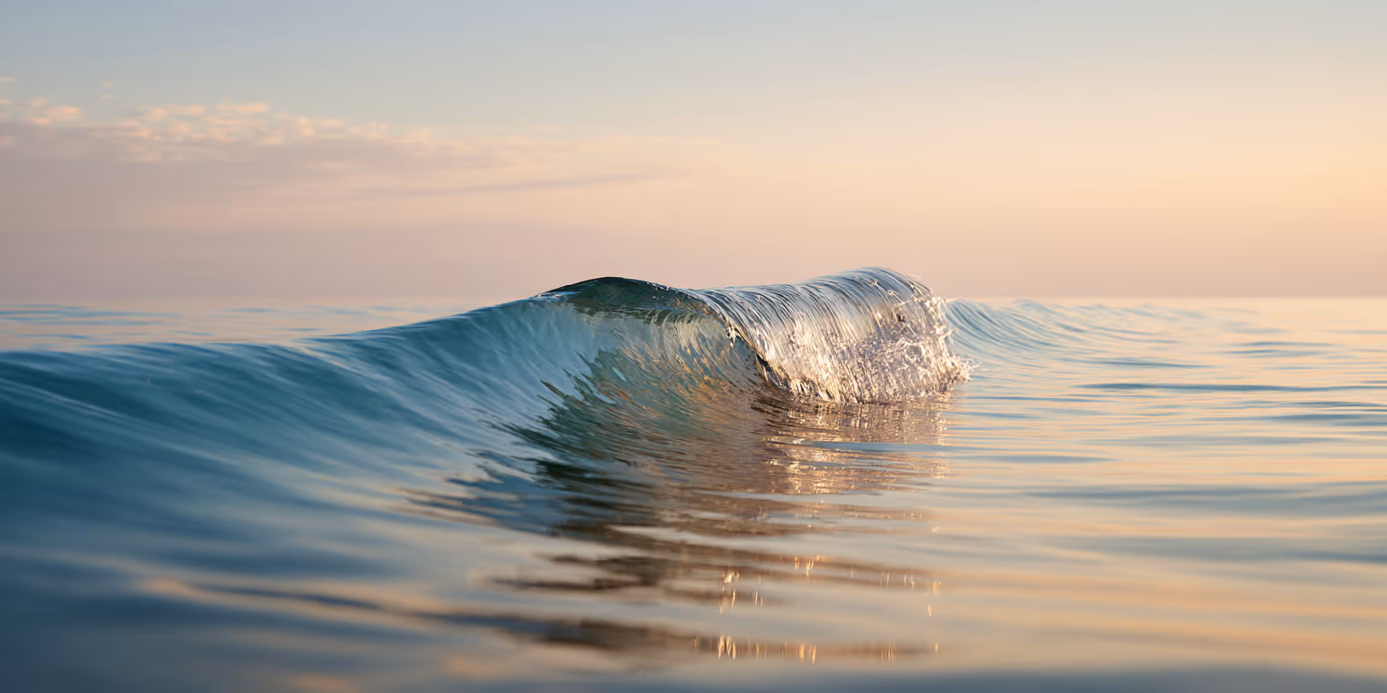 Gentle ocean wave rising under warm sunrise light, reflecting the sky in soft blue and golden tones.