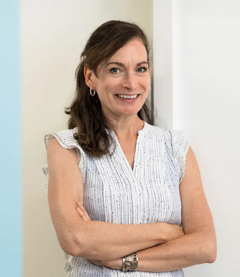 Lächelnde Frau mit dunklen Haaren, die mit verschränkten Armen vor heller Wand steht, trägt weiße Bluse mit feinen Streifen.