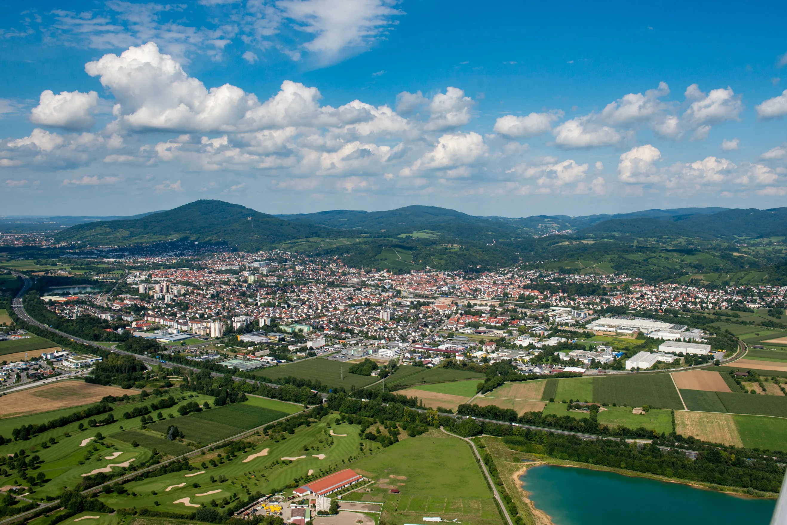 Luftaufnahme einer Stadt mit Häusern, Feldern, einem Golfplatz und einem See vor bewaldeten Hügeln unter blauem Himmel mit Wolken.