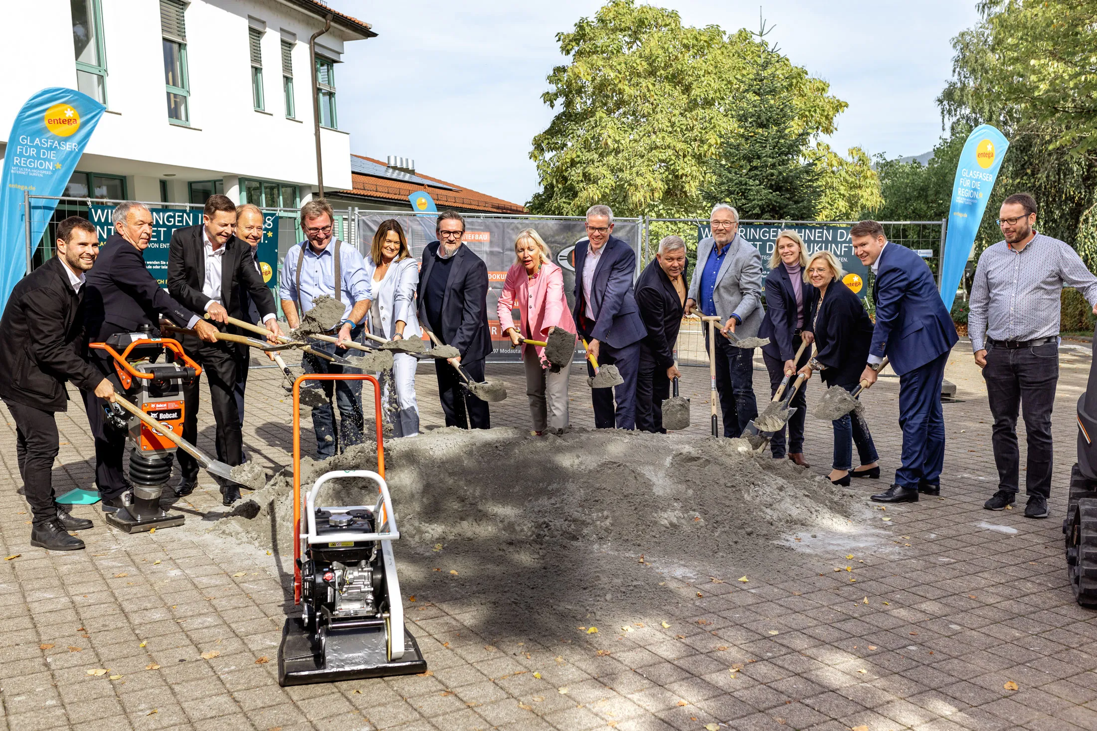 Gruppe von Menschen bei einem feierlichen Spatenstich mit Schaufeln vor einem großen Sand- oder Zementhaufen im Freien.