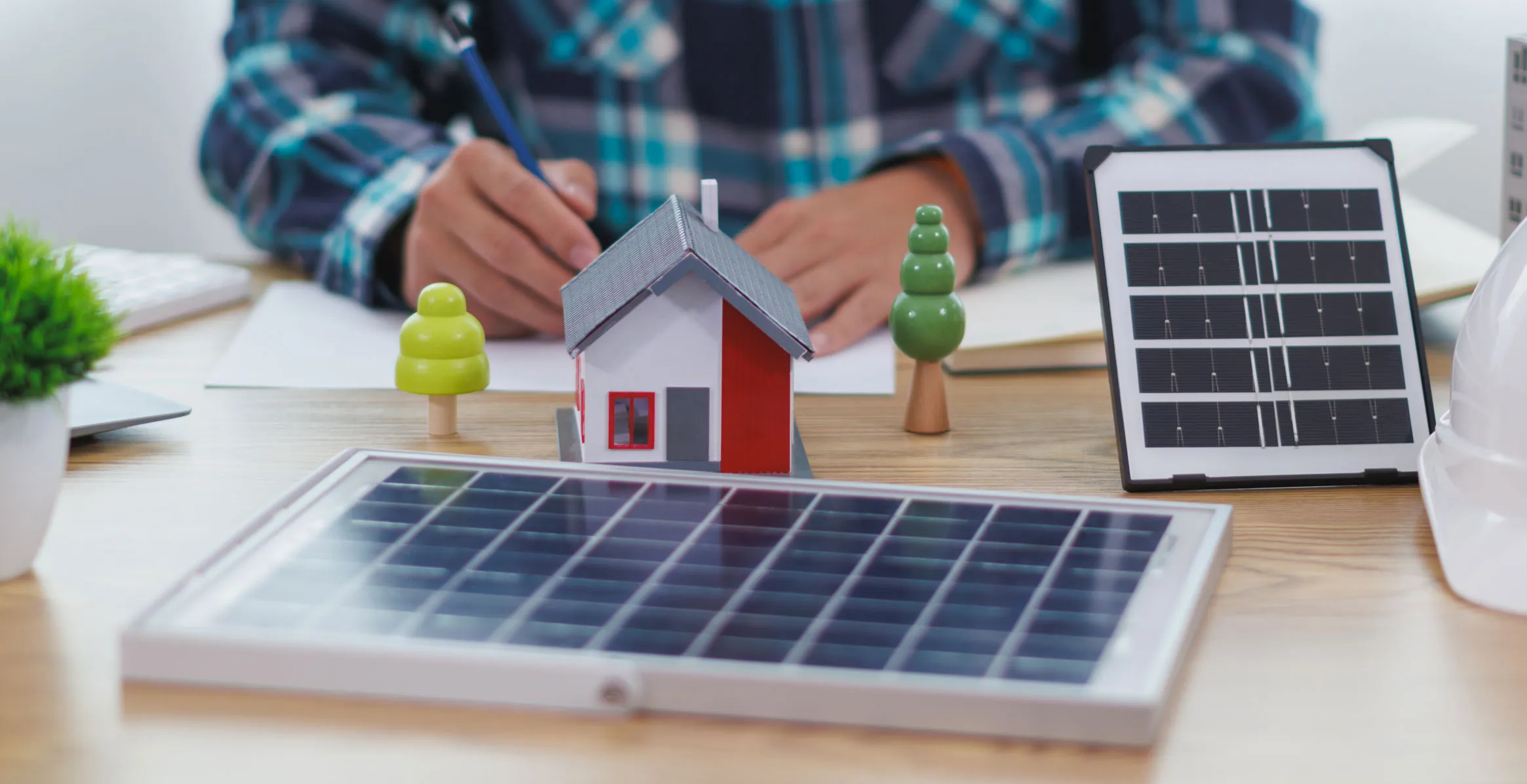 Person schreibt hinter einem Modellhaus, zwei Miniaturbäumen, zwei Solarpanels und einem weißen Schutzhelm auf einem Schreibtisch.