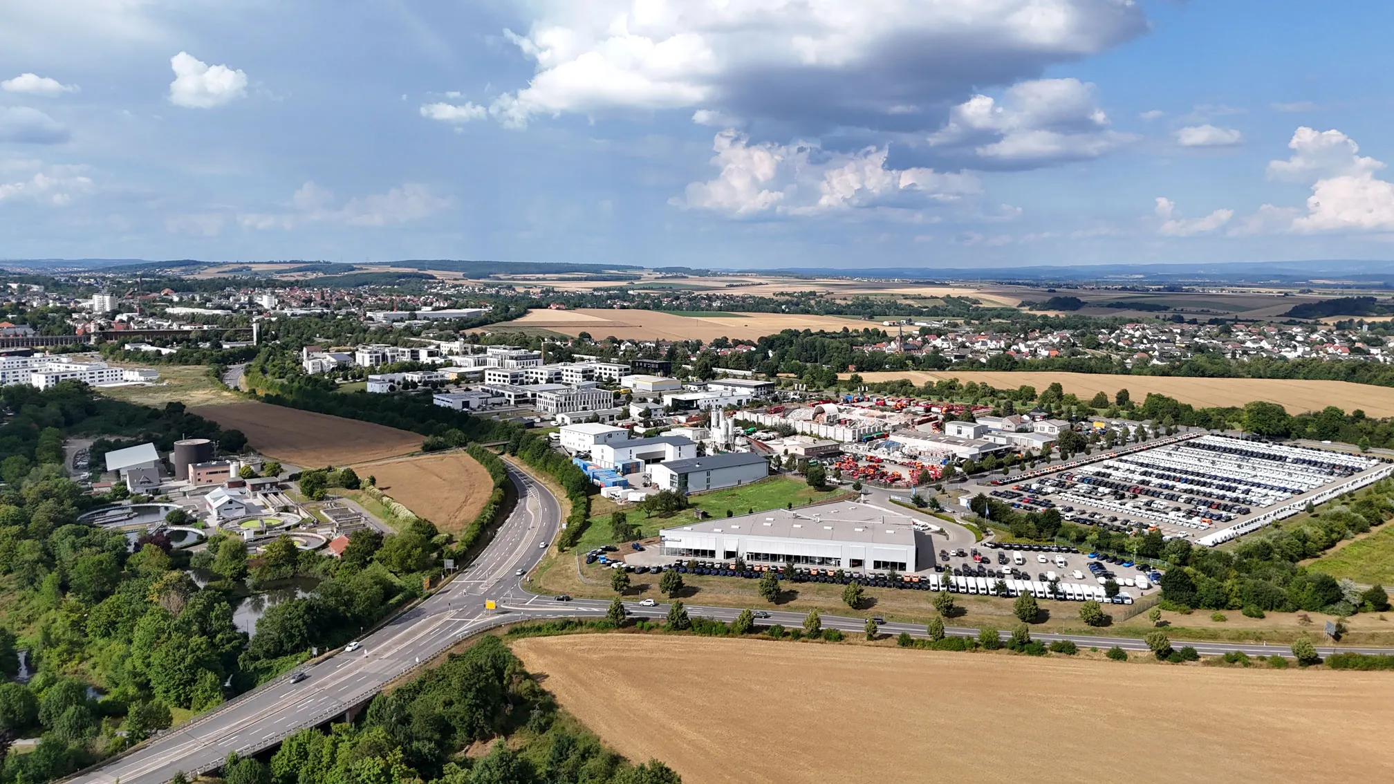 Luftaufnahme eines Gewerbegebiets mit mehreren Gebäuden, Parkplätzen voller Fahrzeuge und umgebenden Feldern unter blauem Himmel mit Wolken.