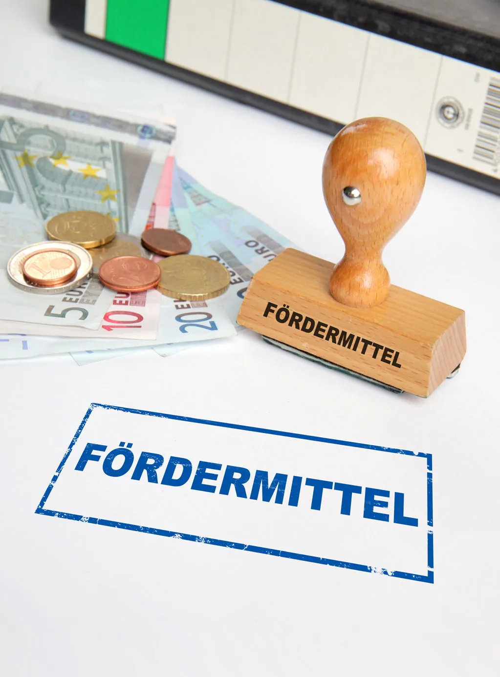 Holzstempel mit der Aufschrift ‚FÖRDERMITTEL‘ neben Euro-Geldscheinen und Münzen auf weißem Untergrund.
