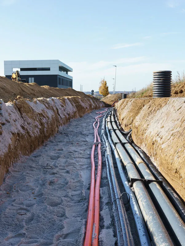 Kabelverlegung im offenen Graben mit mehreren schwarzen und roten Rohren neben einem modernen Gebäude bei klarem Himmel.